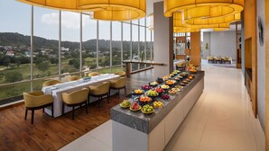 Un moderno comedor con una larga barra llena de frutas y bebidas, ventanas amplias que ofrecen vistas al campo, e iluminación amarilla cálida.