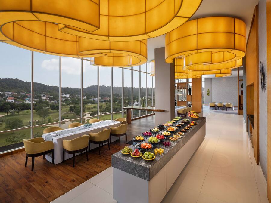 A modern dining area with a long counter filled with fruits and beverages, large windows offering a view of the countryside, and warm yellow lighting.