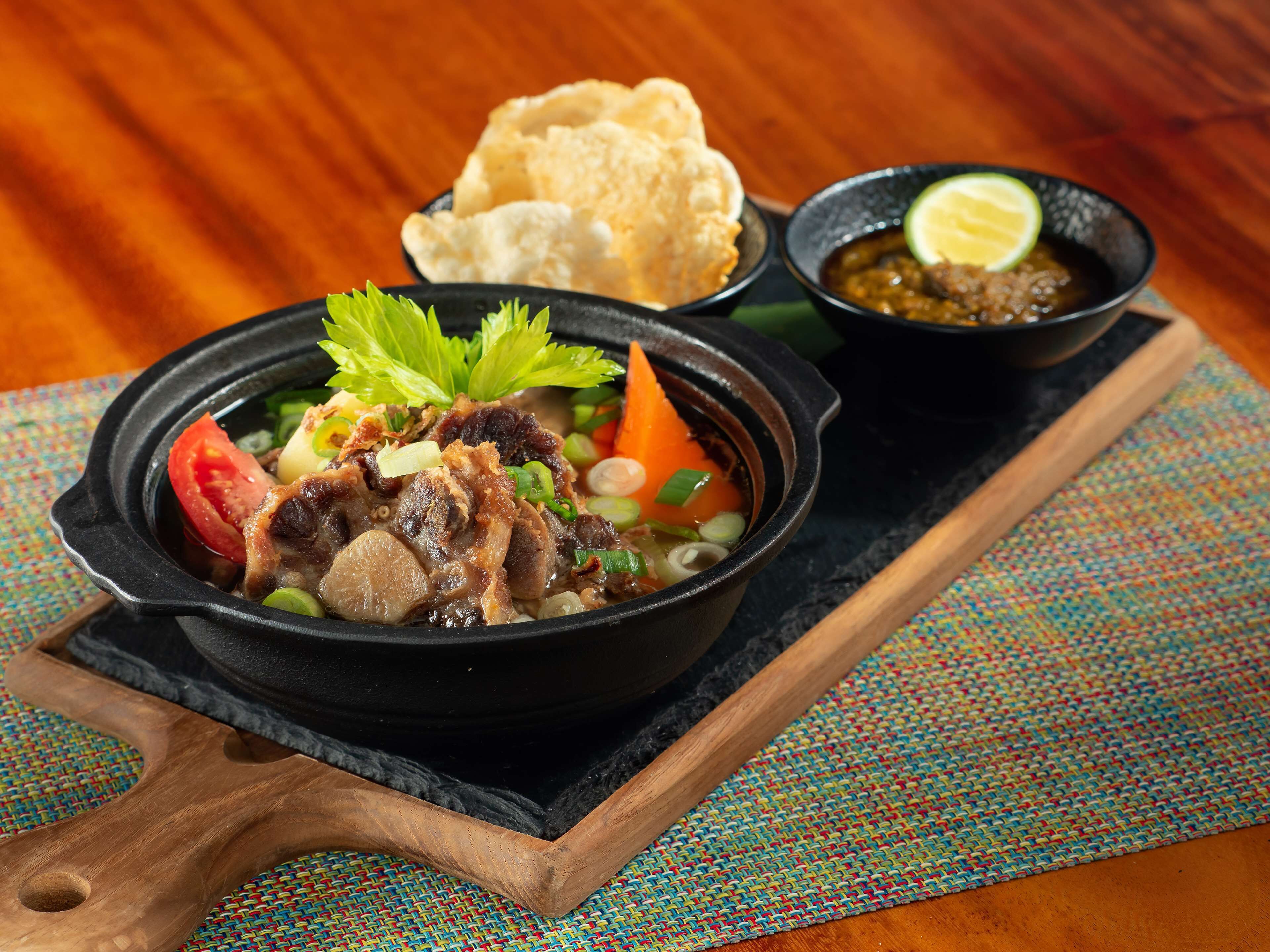 A bowl of soup with vegetables and meat, accompanied by a side of flatbread and a small bowl of sauce on a wooden board.