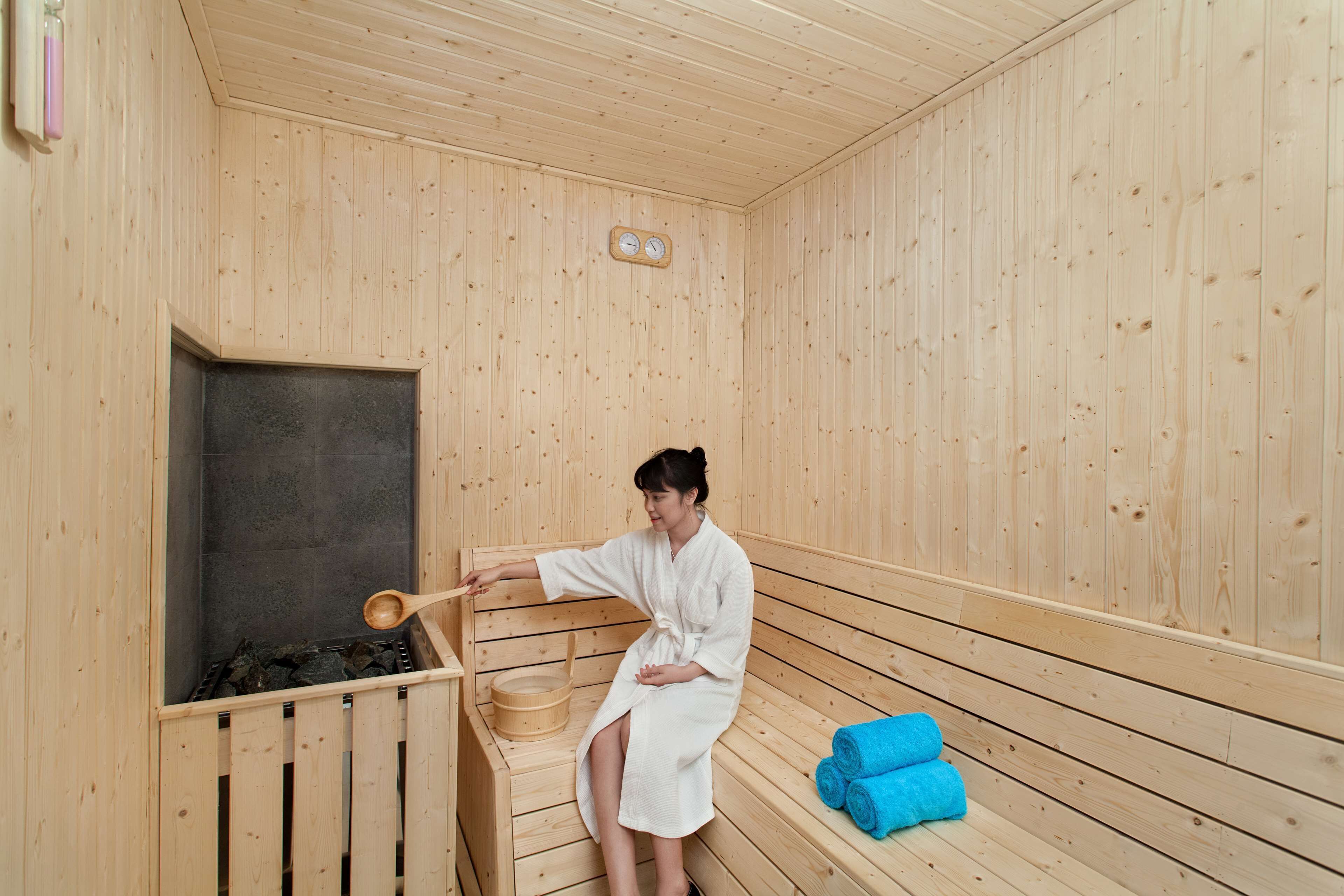 A wooden sauna with a person in a white robe, a wooden bench, and a blue towel on the floor.