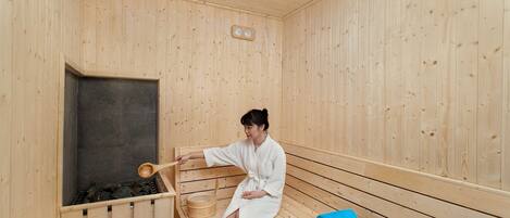 A wooden sauna with a person in a white robe, a wooden bench, and a blue towel on the floor.