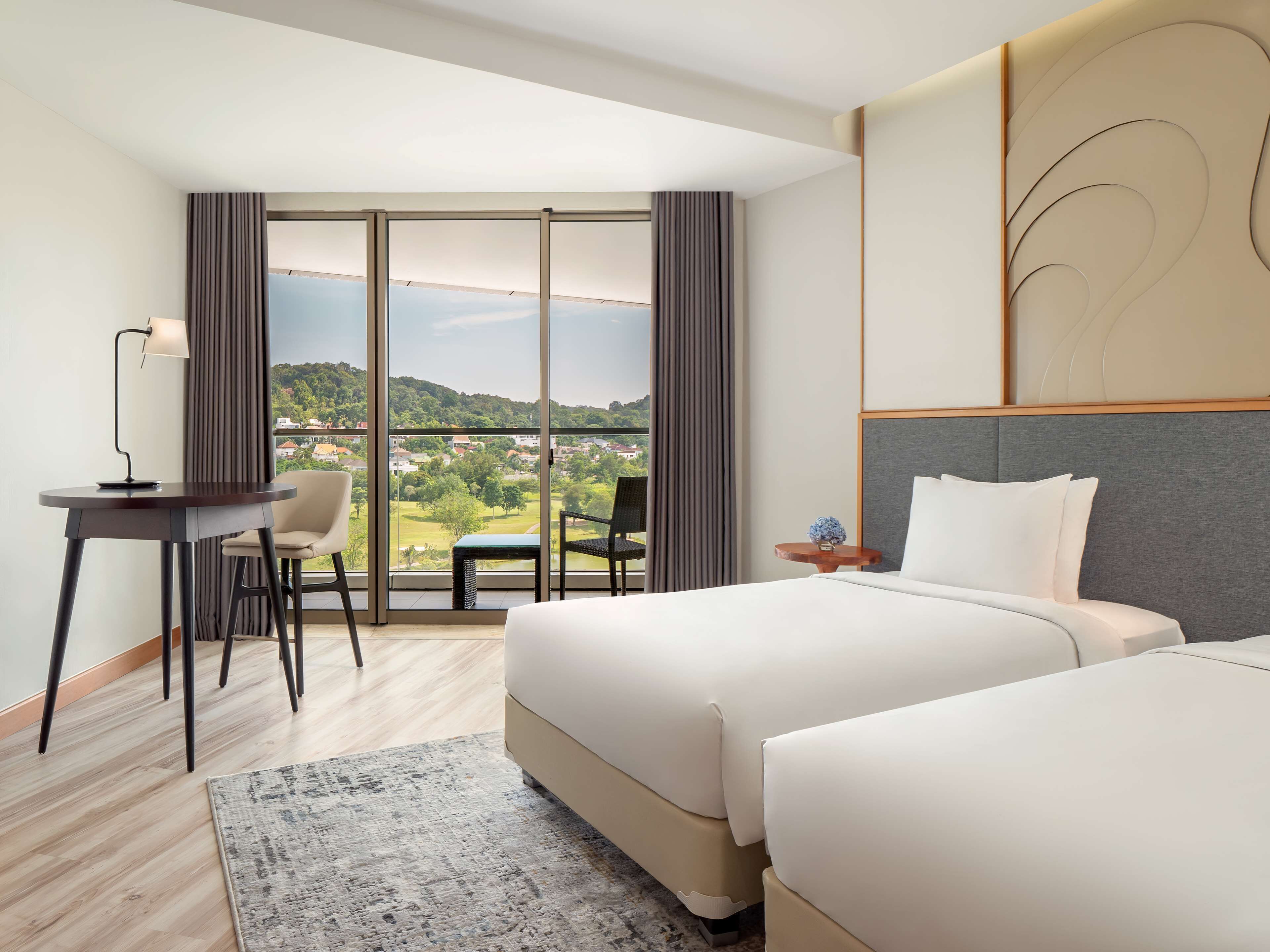 A hotel room with a large bed, a desk with a chair, a small table with a vase, and a view of a green landscape through large windows.