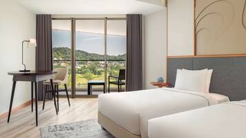 A hotel room with a large bed, a desk with a chair, a small table with a vase, and a view of a green landscape through large windows.