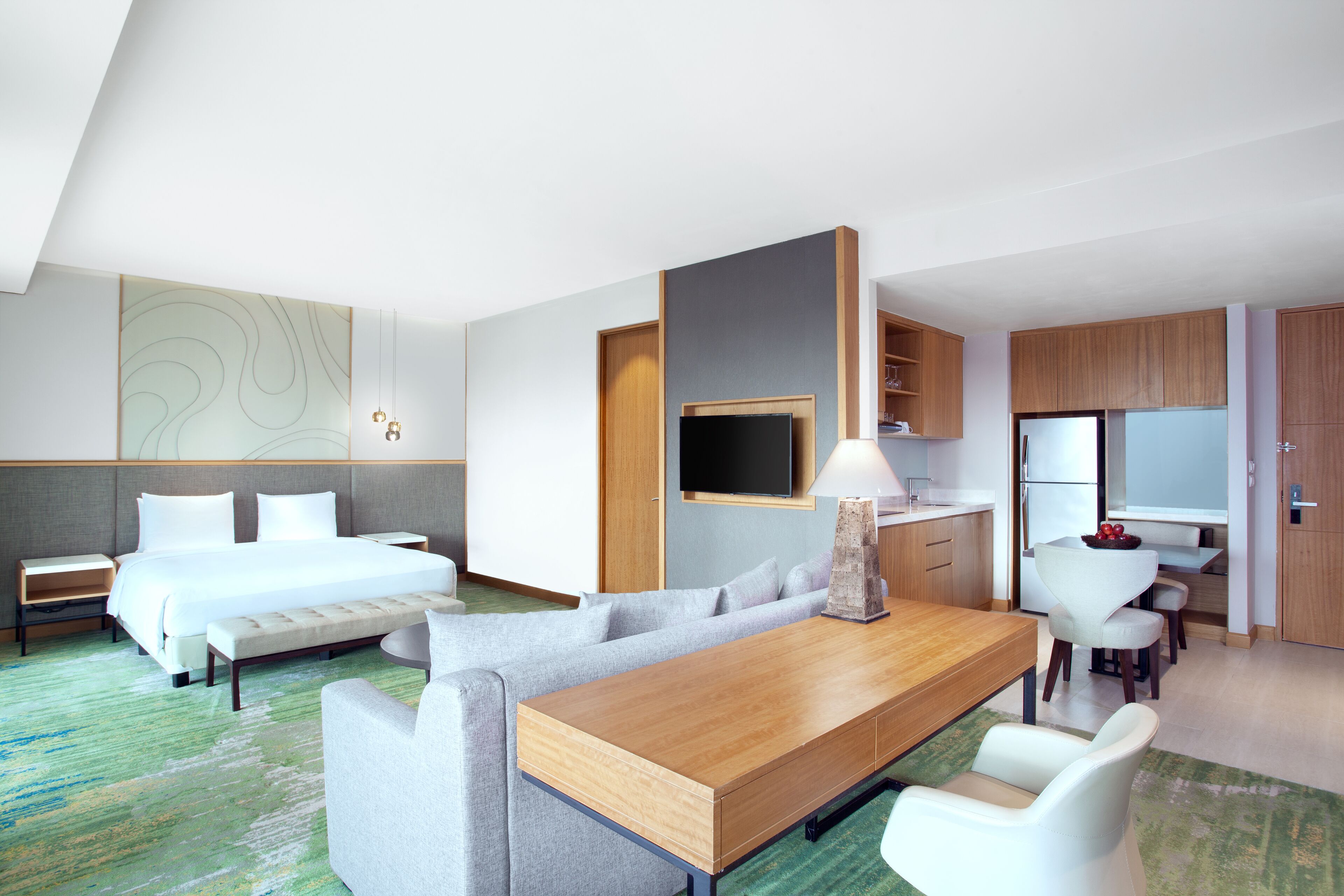 A modern hotel room with a bed, a sofa, a dining table, a TV, and a kitchenette.