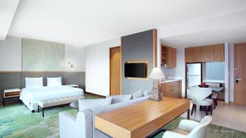 A modern hotel room with a bed, a sofa, a dining table, a TV, and a kitchenette.