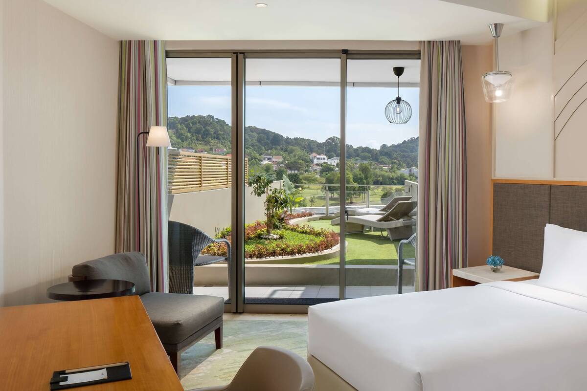 Ein Hotelzimmer mit einem großen Bett, einem Schreibtisch, einem Sessel und Blick auf einen angelegten Garten sowie Gebäude in der Ferne.