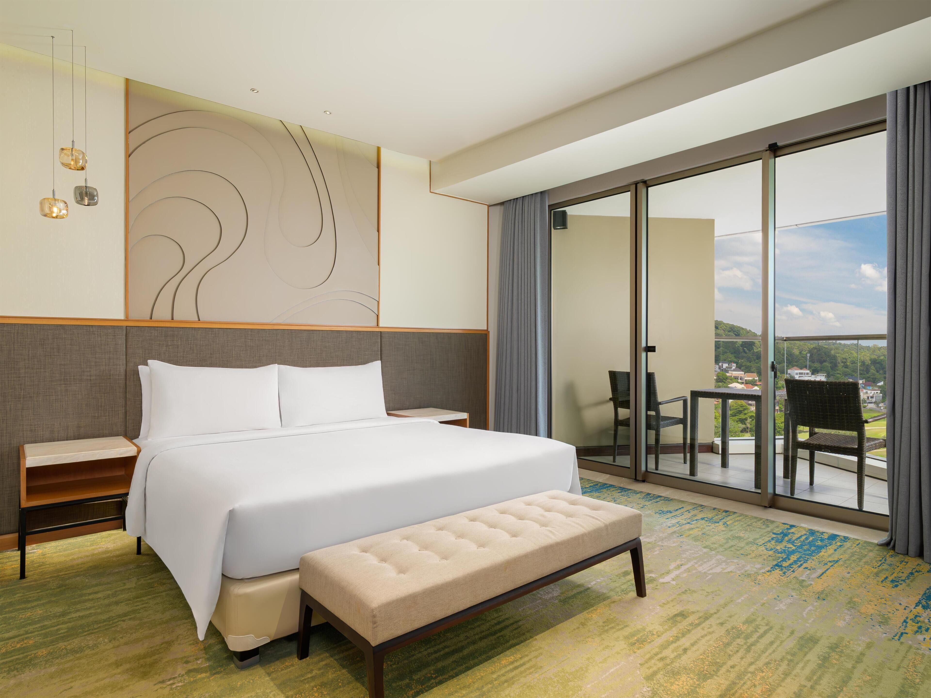 A hotel room with a large bed, a bench, a nightstand, a wall-mounted light fixture, and a sliding glass door leading to a balcony with chairs.