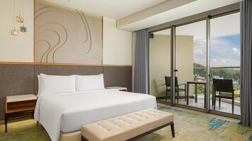 A hotel room with a large bed, a bench, a nightstand, a wall-mounted light fixture, and a sliding glass door leading to a balcony with chairs.