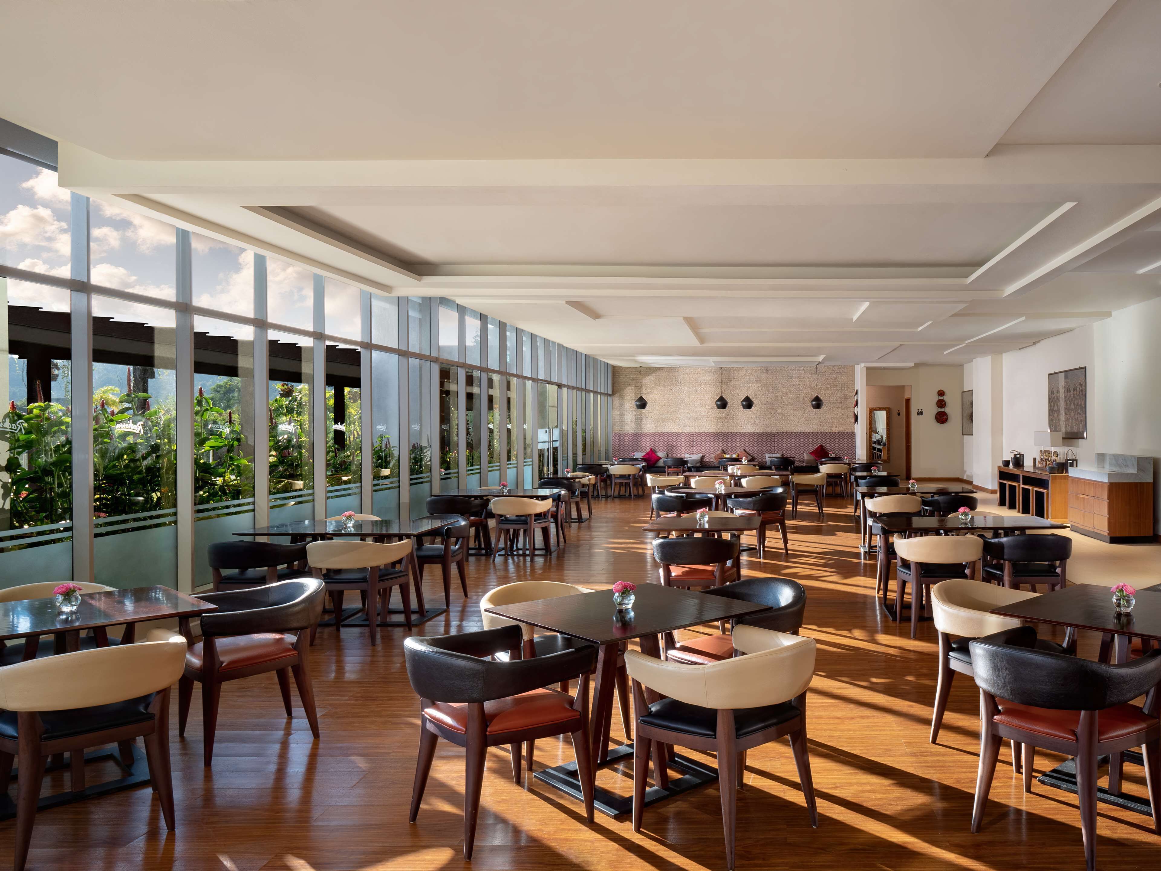Un vaste espace dédié à la salle à manger, avec de nombreuses tables et chaises, de grandes fenêtres offrant une vue sur la verdure et un aménagement intérieur moderne.