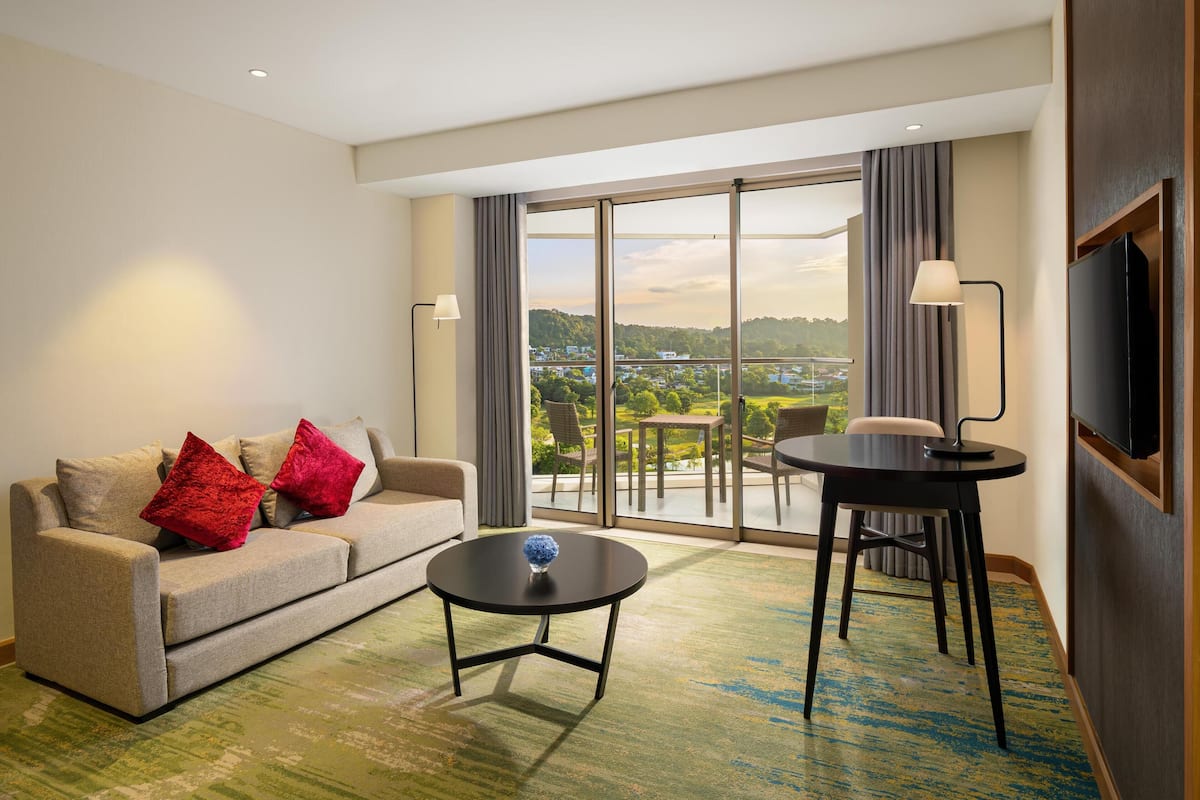 Ein modernes Hotelzimmer mit einer Couch, einem kleinen Tisch, einem Sessel, einem Fernseher und einem Balkon mit Blick auf die Landschaft.