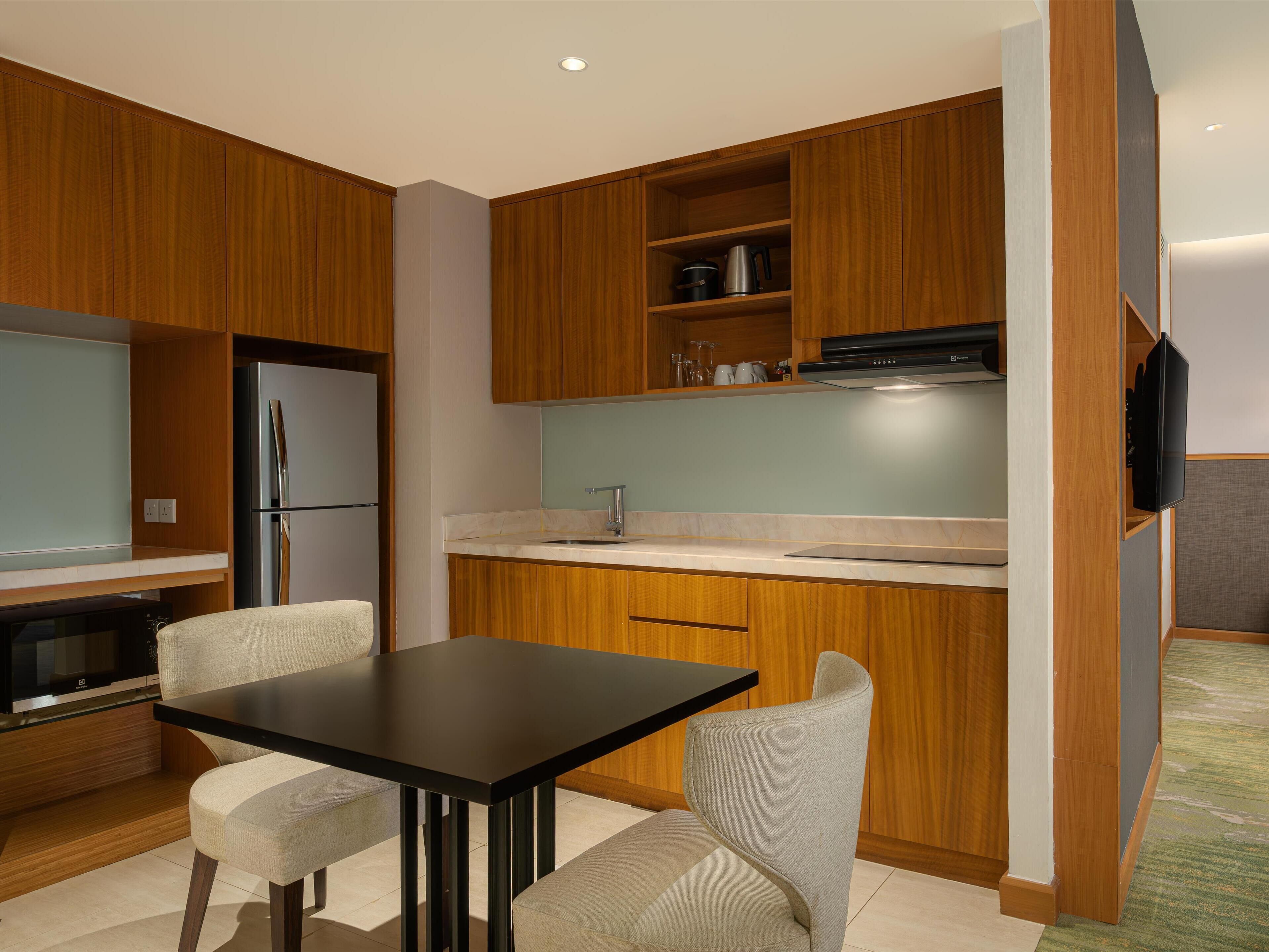 Cocina moderna con armarios de madera, encimera negra, refrigerador de acero inoxidable, horno integrado y televisor de pantalla plana montado en la pared.