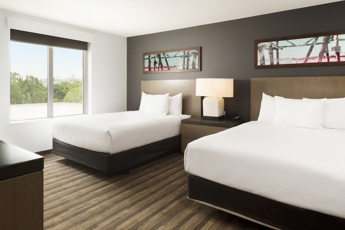 A hotel room with two beds, a large window, a nightstand with a lamp, and two framed pictures on the wall.
