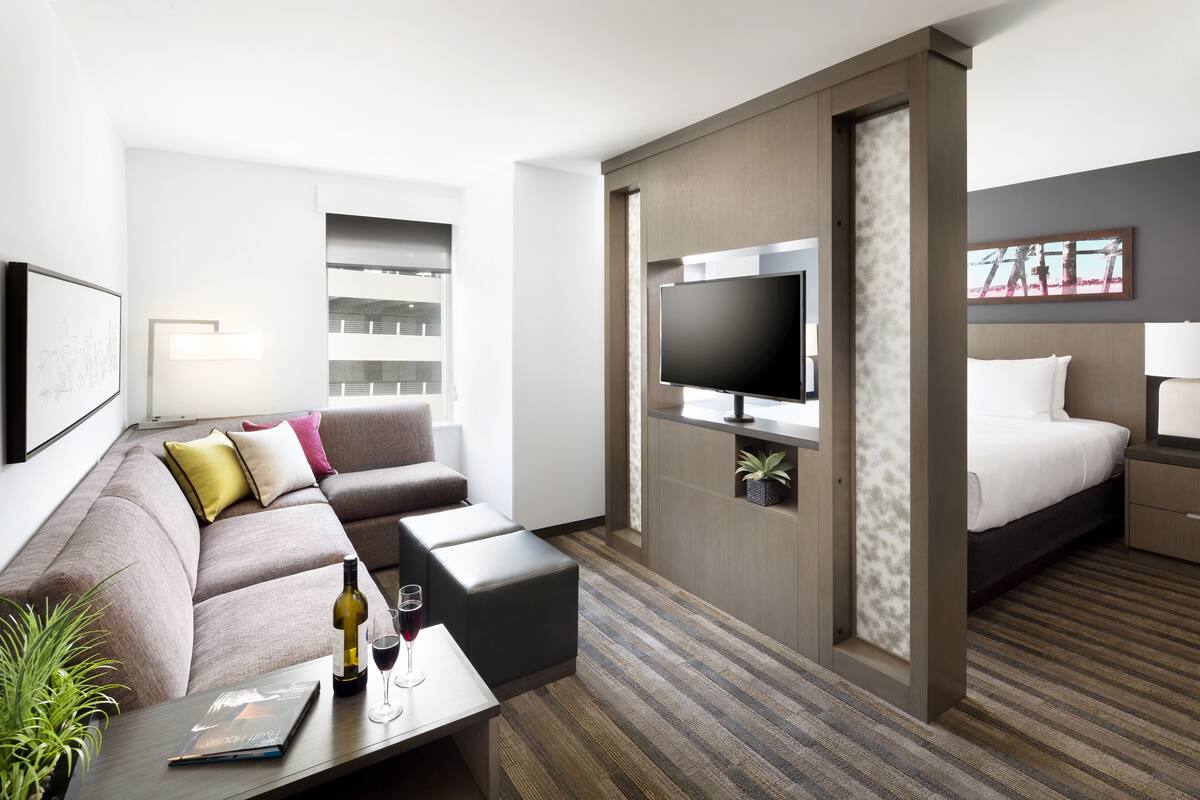 A modern hotel room with a sofa, a flat-screen TV, a bed, and a small table with a wine bottle and glasses.