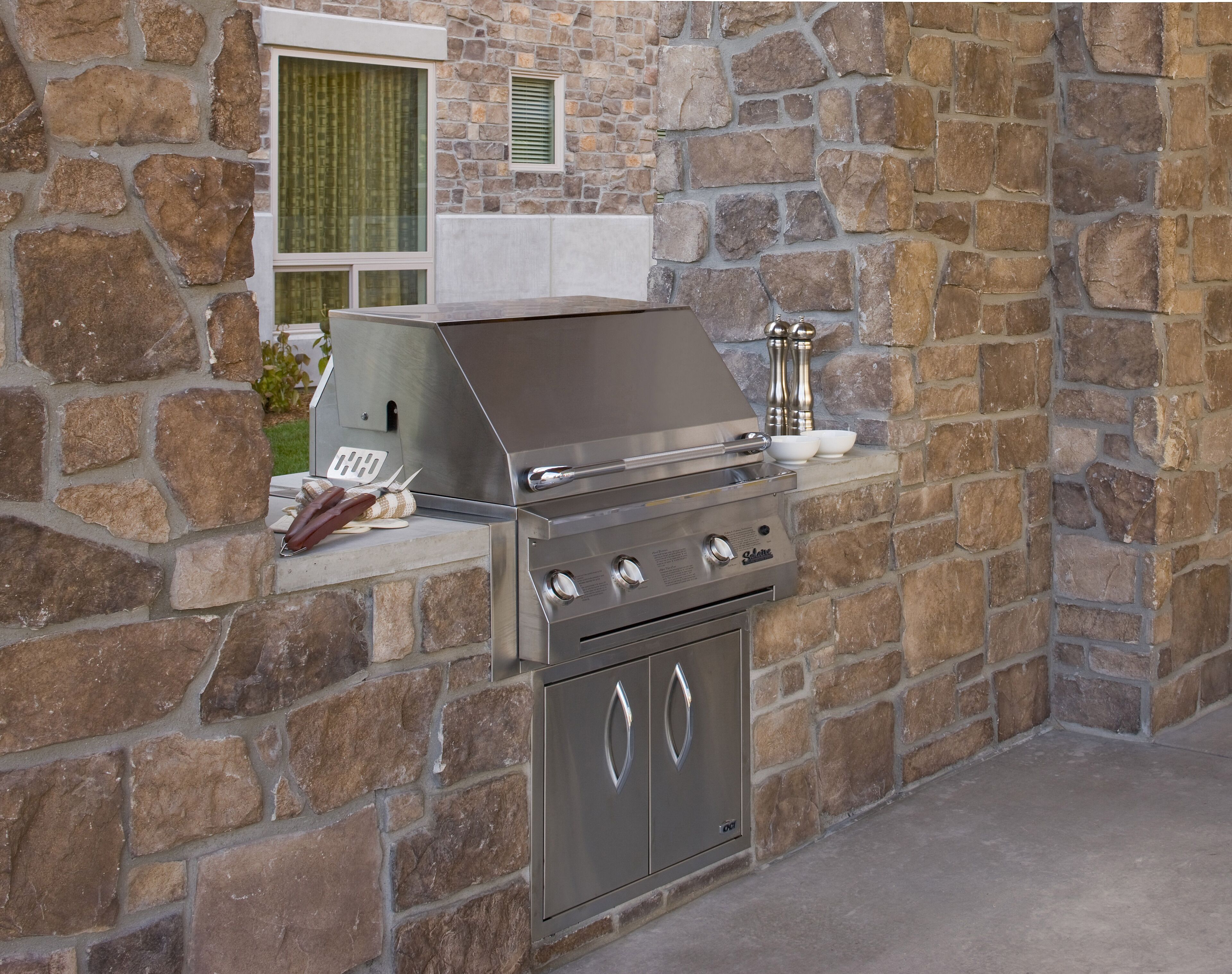 A stainless steel grill with a built-in oven, set against a stone wall.