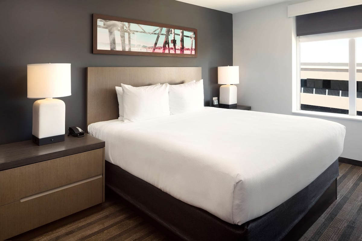 A hotel room with a large bed, two bedside tables with lamps, a nightstand, a painting on the wall, and a window with a view.