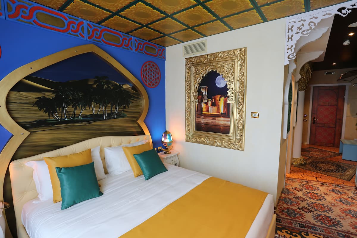 Una habitación de hotel con una cama grande, una mesita de noche con lámpara, un muro decorativo con luna y estrellas, y una alfombra estampada.