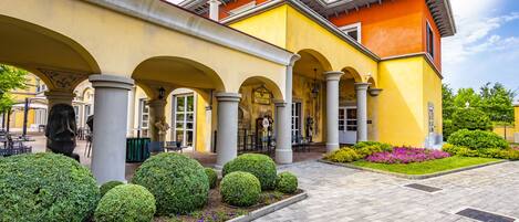 A building with a yellow facade, multiple arches, and a paved courtyard with neatly trimmed bushes and flowers.
