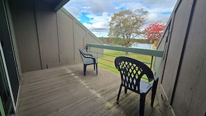 Una terraza de madera con dos sillas y una mesa, con vista a un área verde y un lago.