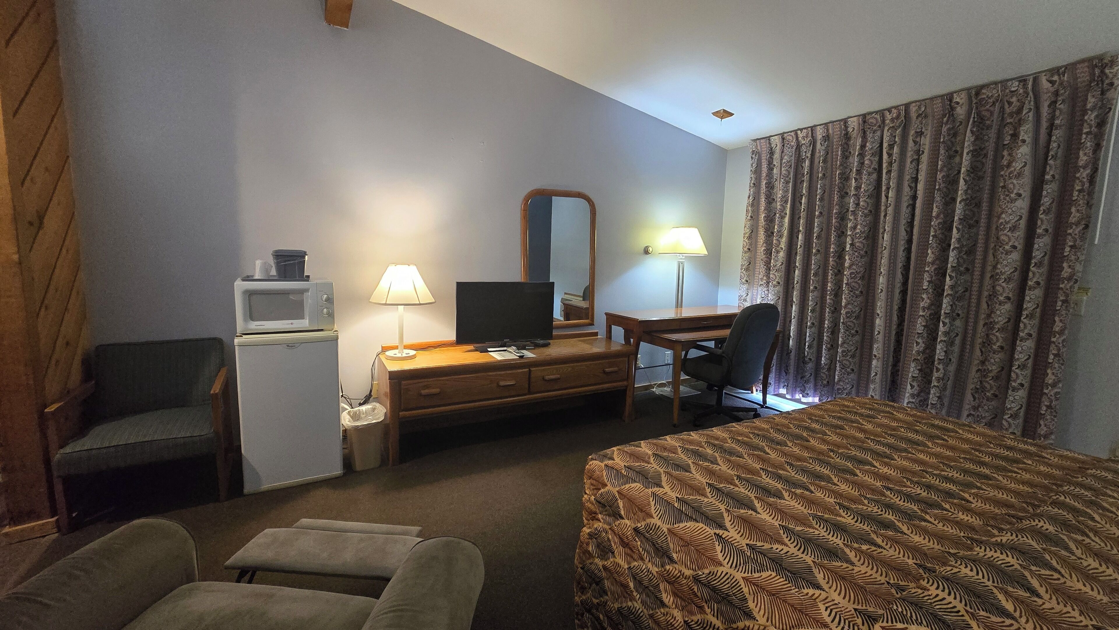 Ein Hotelzimmer mit einem Bett, einem Schreibtisch mit Lampe, einem Fernseher, einer Mikrowelle, einem Stuhl und einem Papierkorb.