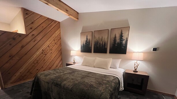 Un dormitorio con techo inclinado de madera, una cama con colcha estampada, dos mesitas de noche con lámparas y tres cuadros en la pared.