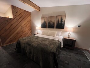 A bedroom with a wooden sloped ceiling, a bed with a patterned bedspread, two bedside tables with lamps, and three framed pictures on the wall.