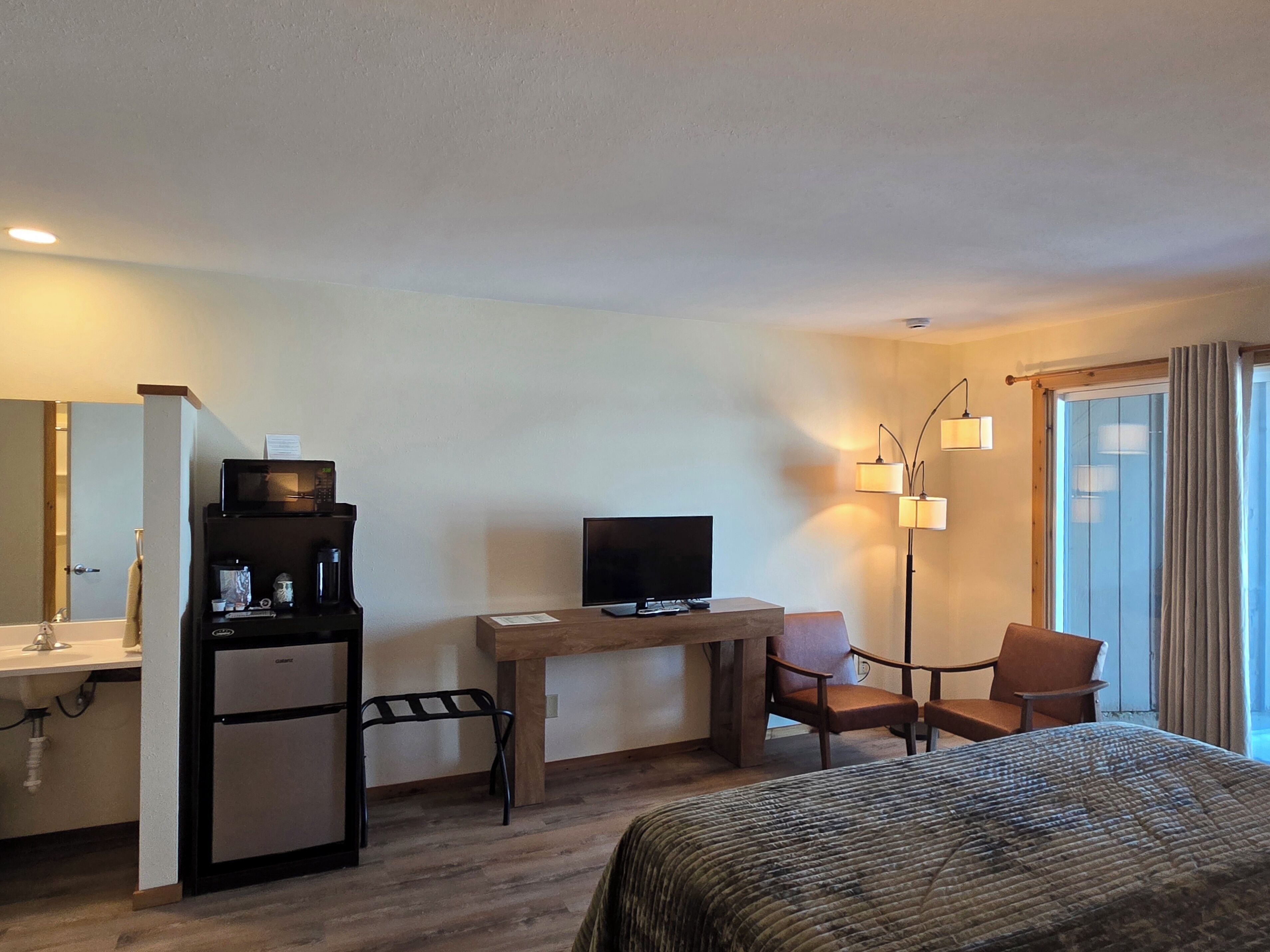 A hotel room with a bed, a desk with a television, a refrigerator, a chair, a lamp, and a window with curtains.