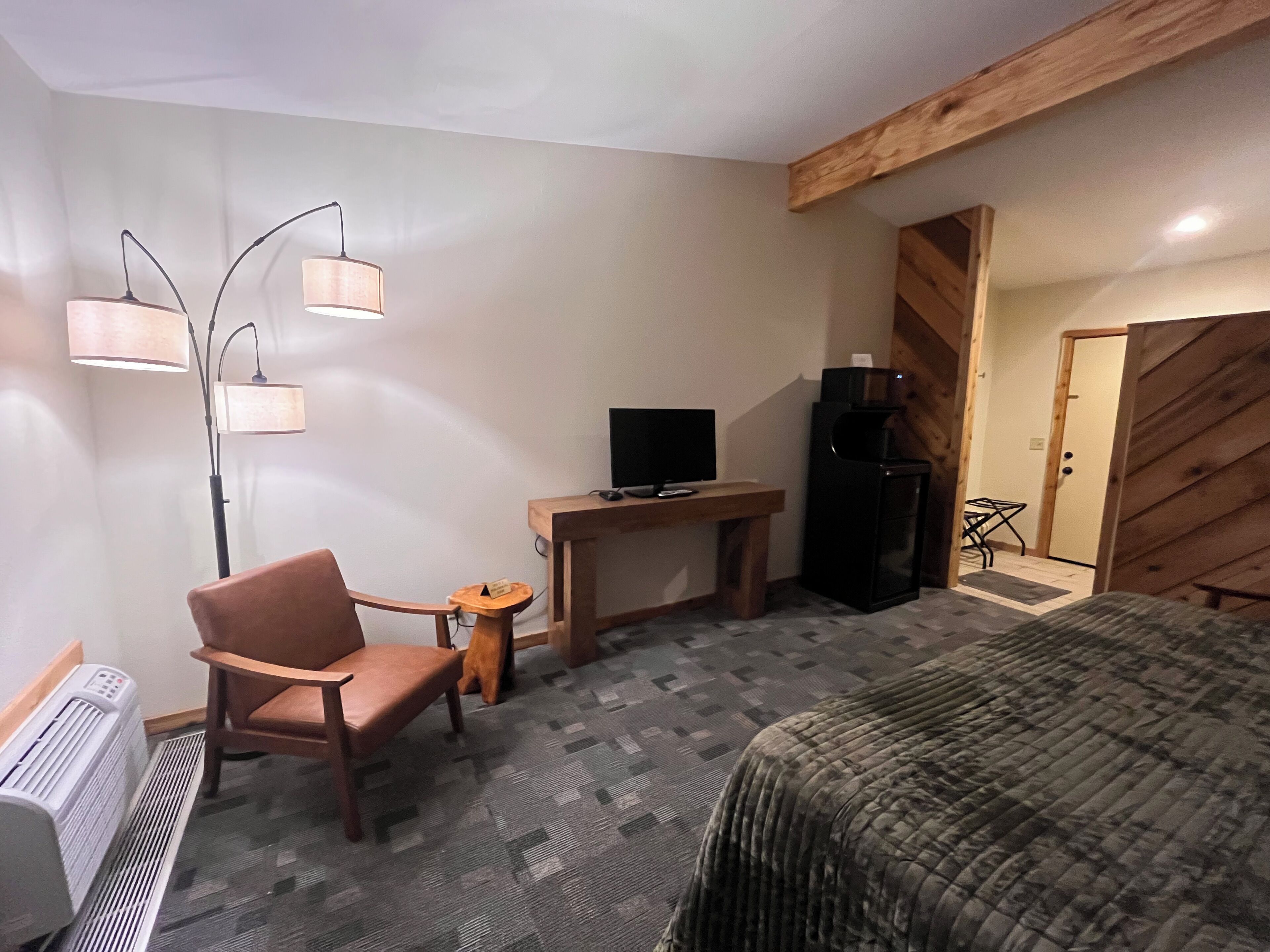 A hotel room with a bed, a desk with a computer, a chair, a standing lamp, a heater, and a refrigerator.