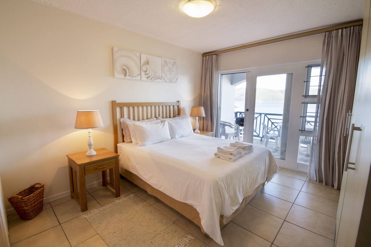 A hotel room with a bed, two bedside tables with lamps, a small basket, and a view of the outside through a sliding glass door.