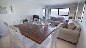 Amplia sala de estar con mesa de comedor de madera y sillas blancas, sofá beige con cojines, televisor de pantalla plana y ventanas grandes que ofrecen vistas al agua y las colinas.