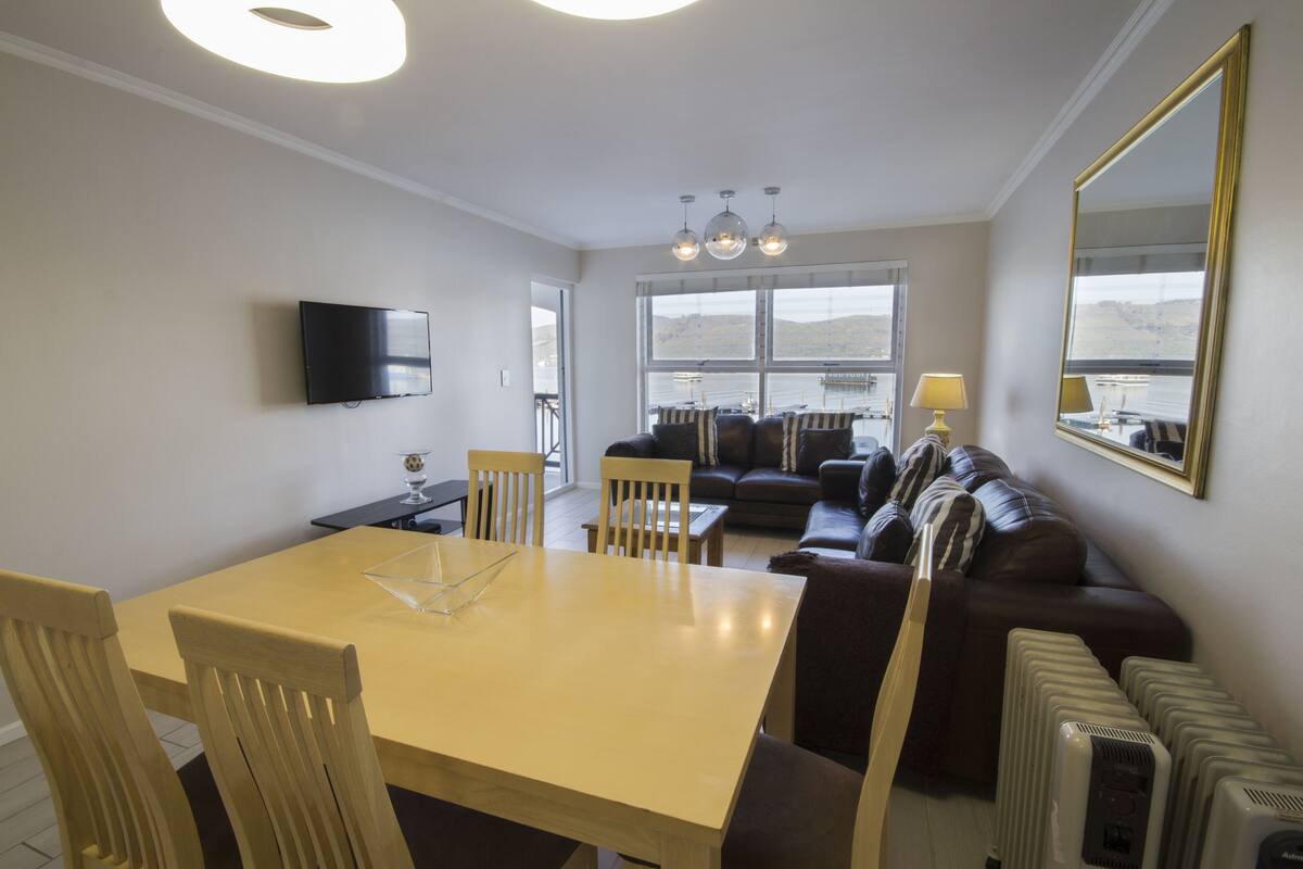 A modern living room with a dining area, a sofa set, a television mounted on the wall, and a large window offering a view of the outdoors.
