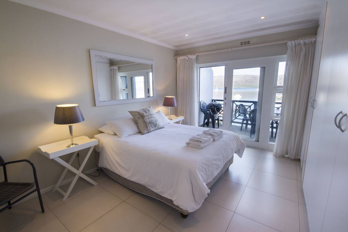A bedroom with a bed, bedside tables, a chair, a mirror, and a view of an outdoor area with chairs and a table.