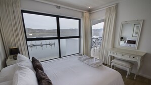 Habitación de hotel con un ventanal que ofrece vistas a un cuerpo de agua y colinas lejanas. Hay una cama con ropa blanca y un banco al lado.