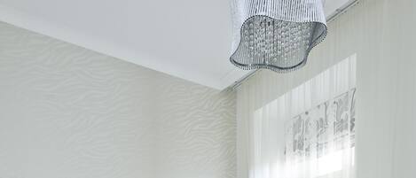 Una cama grande con cabecera capitonada, ropa de cama blanca y almohadas estampadas. Hay una mesita de noche con una lámpara, y una ventana con cortinas translúcidas.