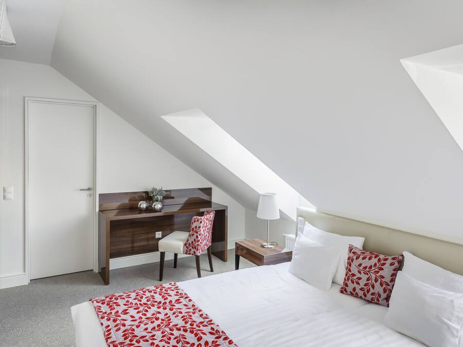 Un dormitorio con techo inclinado, cama con ropa blanca y almohadas estampadas con hojas, escritorio con silla y armario de madera.