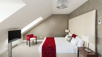 Habitación de hotel moderna con techo inclinado, una cama grande con ropa blanca y un plaid rojo, una silla roja, una mesita de noche con lámpara y un televisor de pantalla plana montado en la pared.