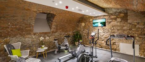 A gym with a brick wall, a treadmill, an elliptical, a stationary bike, and a chair with a green cushion.