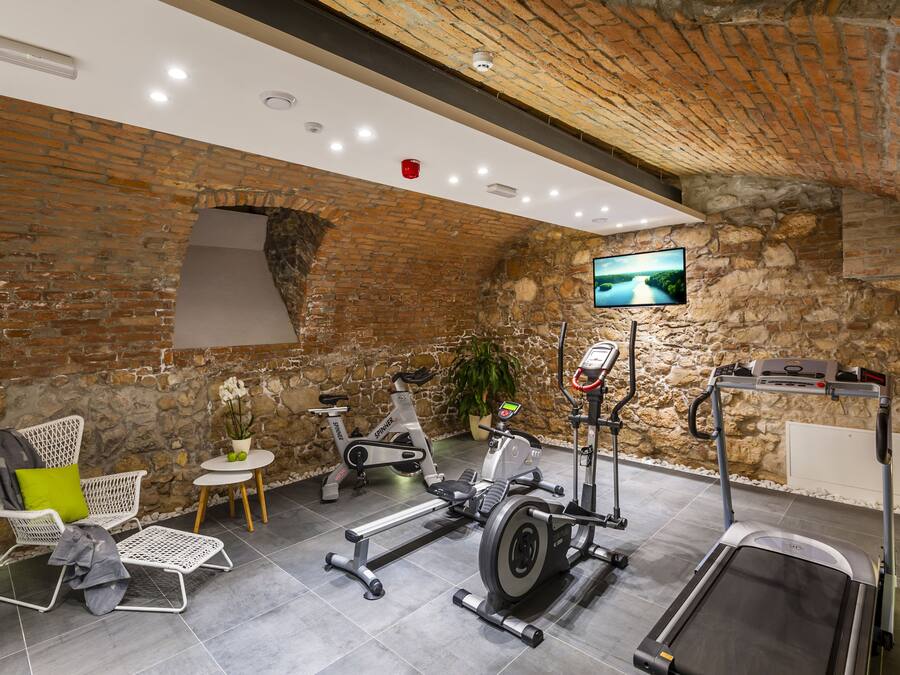 A gym with a brick wall, a treadmill, an elliptical, a stationary bike, and a chair with a green cushion.