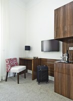 Habitación de hotel con escritorio de madera, una silla, televisor, minibar y maleta.
