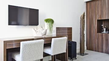 Quarto de hotel com televisão de tela plana na parede, uma mesa com duas cadeiras, uma planta, uma bolsa e uma mala.