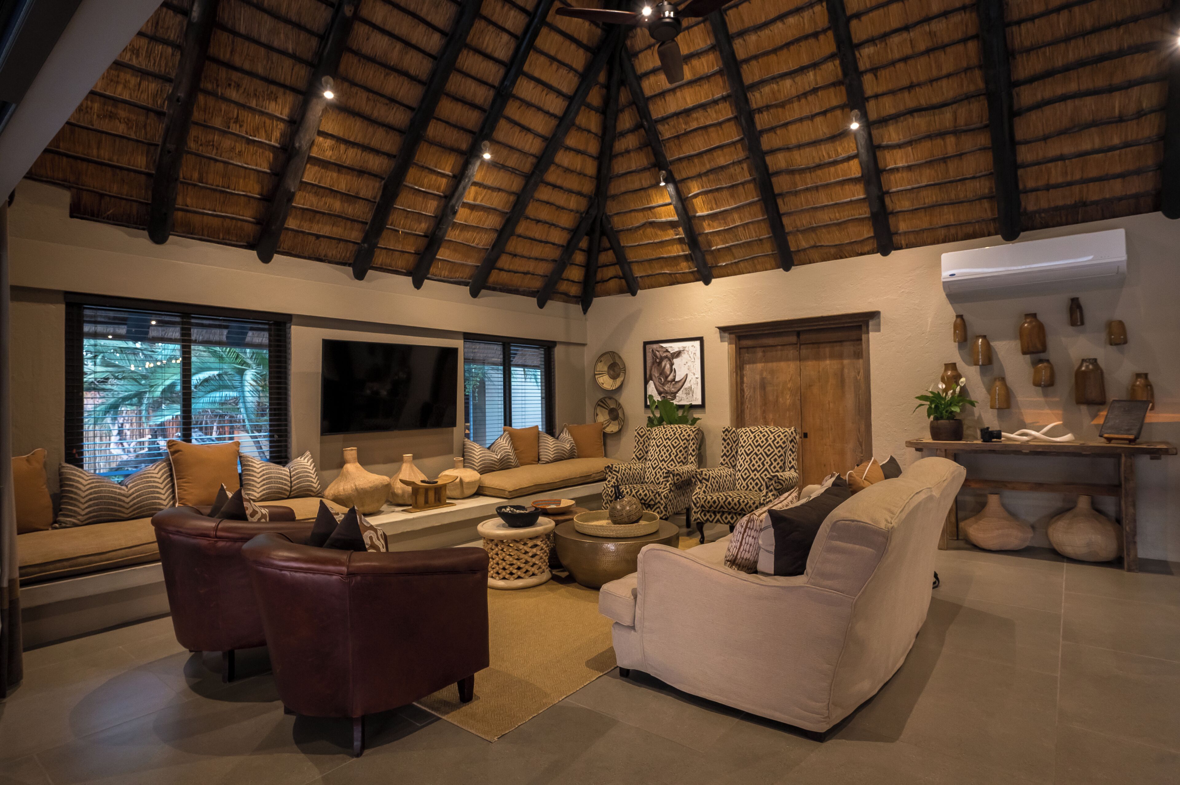 A spacious living room with a high thatched ceiling, a large flat-screen TV, and a variety of seating options including sofas and armchairs.