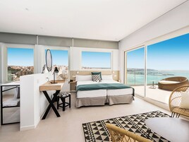 Un dormitorio moderno con cama, escritorio y silla, ventanas amplias con vista a la ciudad y un balcón con sillón de mimbre.