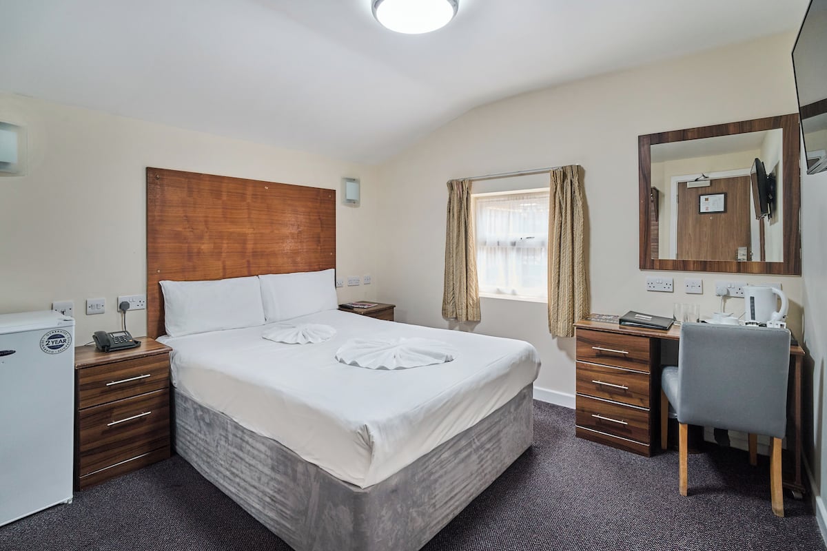 Quarto de hotel com cama, escrivaninha, cadeira, guarda-roupa, espelho e janela com cortinas.