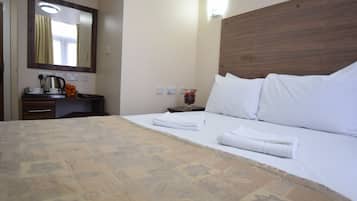 A hotel room with a large bed, a wooden headboard, a nightstand with a lamp, a mirror, and a desk with a coffee maker and flowers.