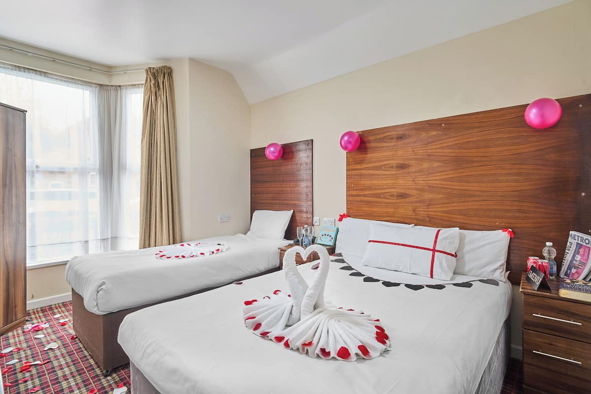 Quarto de hotel com duas camas, uma cabeceira de madeira, uma janela com cortinas e uma mesa de cabeceira com uma revista e uma garrafa.