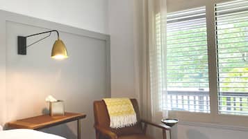 Habitación de hotel con cama, una silla con una manta amarilla, una mesita de noche con lámpara y una ventana con persianas.