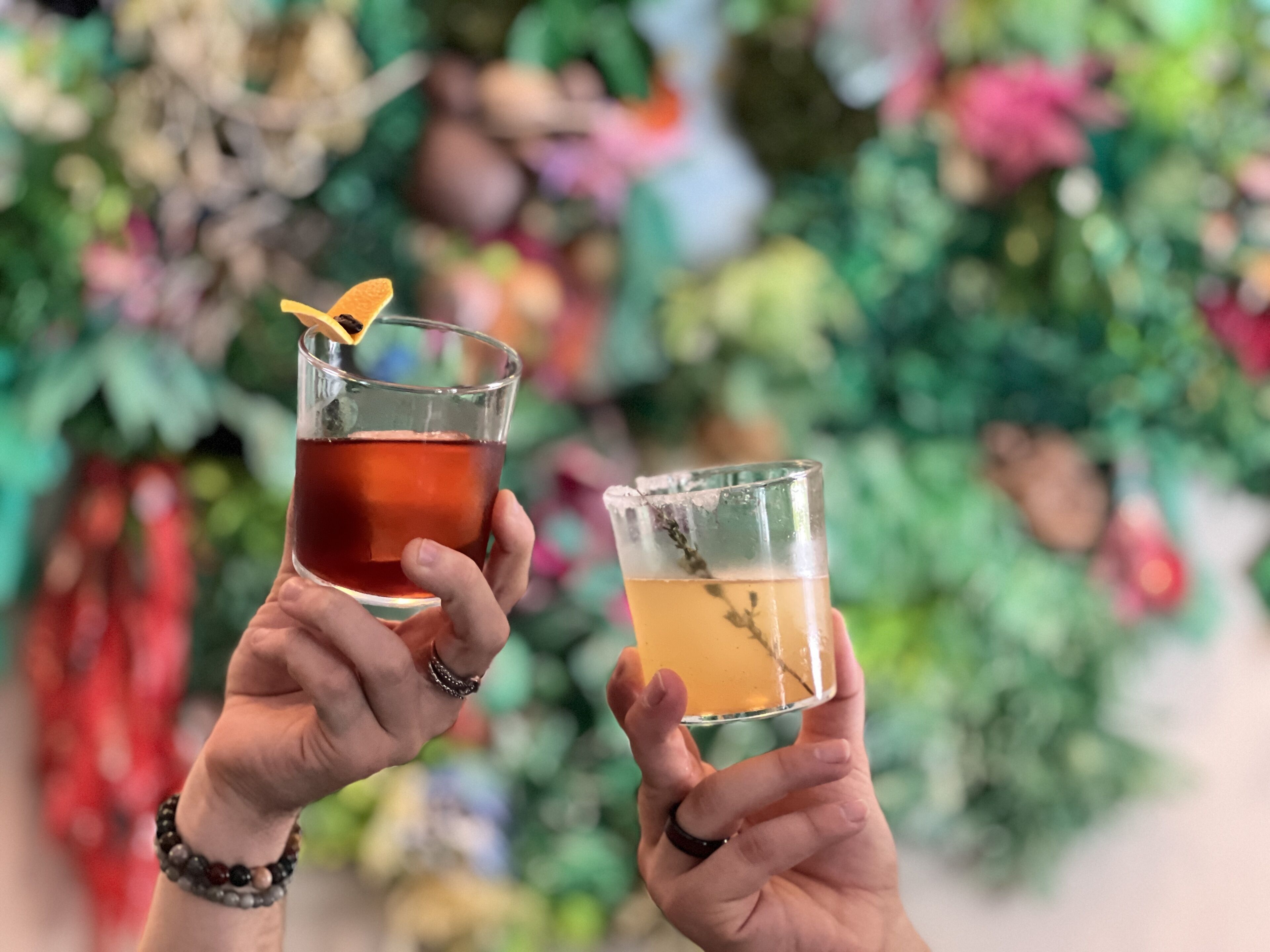 Two people holding glasses of beverages, one with a dark liquid and the other with a light one, in front of a blurred background with greenery.