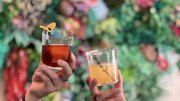 Dos personas sosteniendo vasos con bebidas, uno con líquido oscuro y otro con líquido claro, frente a un fondo borroso con follaje.