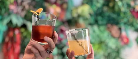 Two people holding glasses of beverages, one with a dark liquid and the other with a light one, in front of a blurred background with greenery.