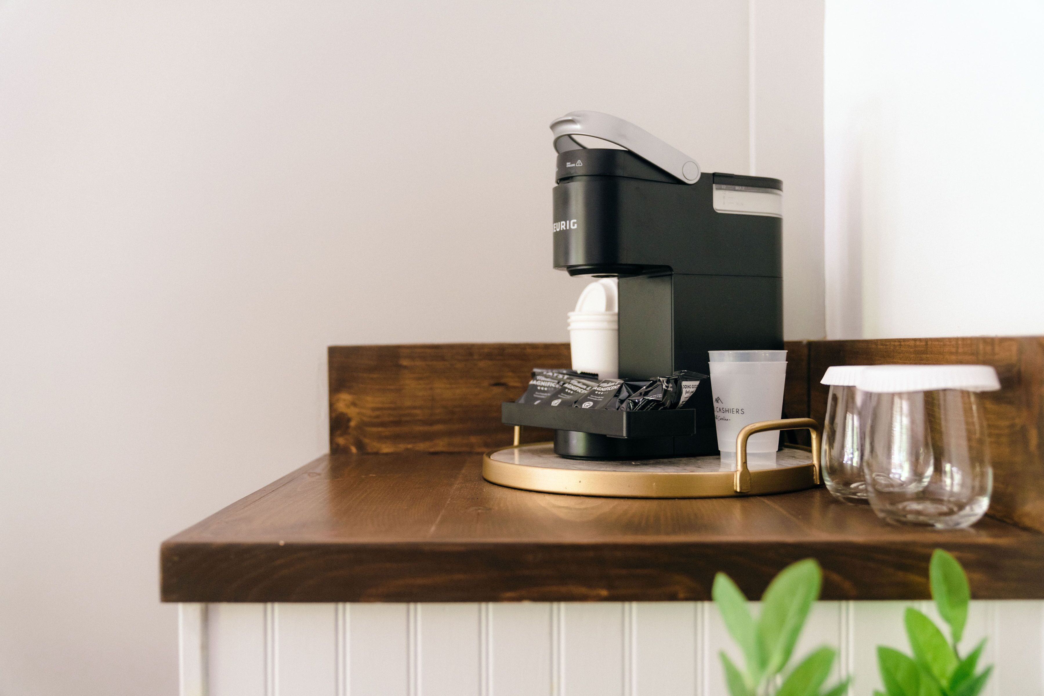 A coffee machine on a wooden countertop with a white panel below, a glass cup, and a small plant.