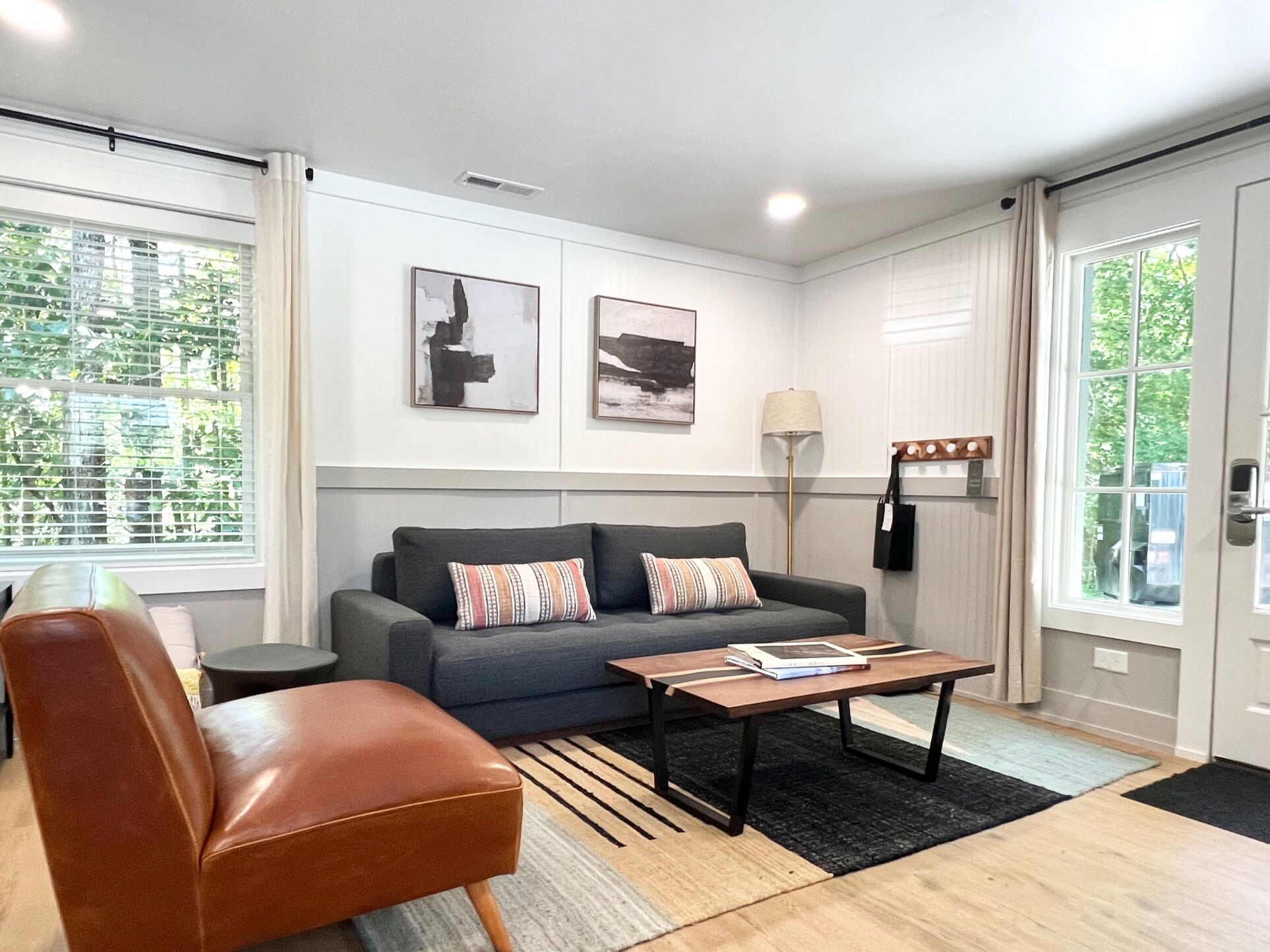 A modern living room with a grey sofa, a brown leather chair, and a coffee table. There are two framed artworks on the wall, a floor lamp, and a window with blinds.